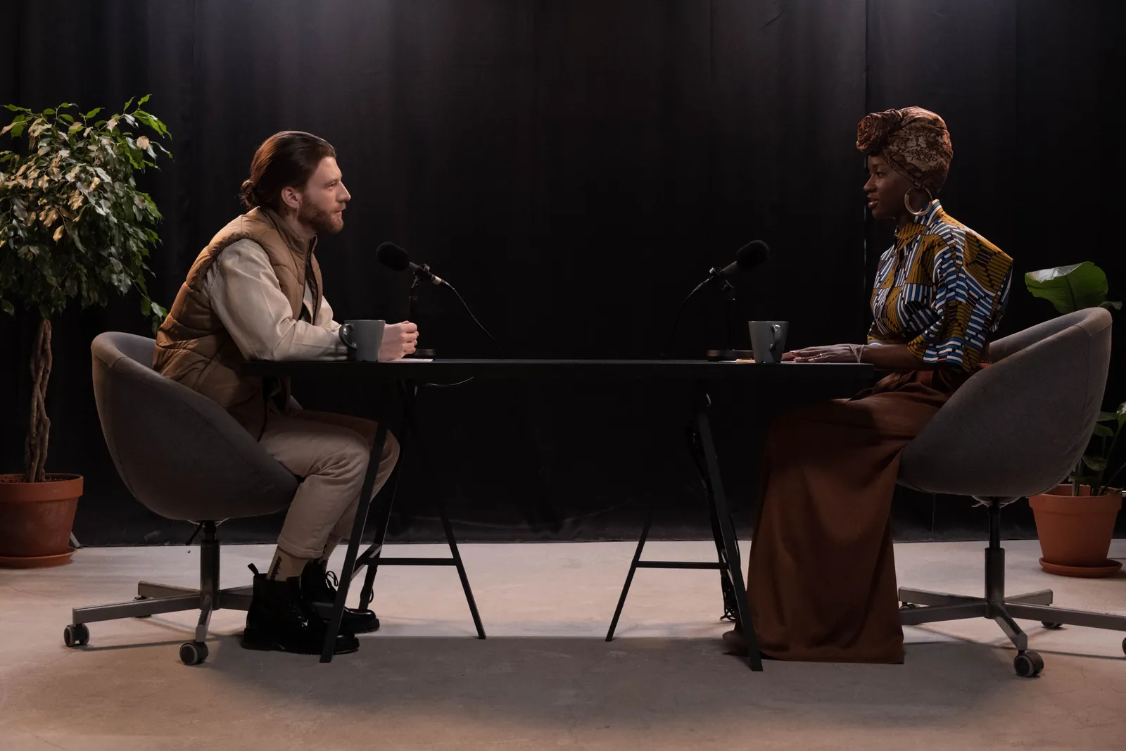 Two podcast hosts facing each other across a table with microphones in a dark studio.