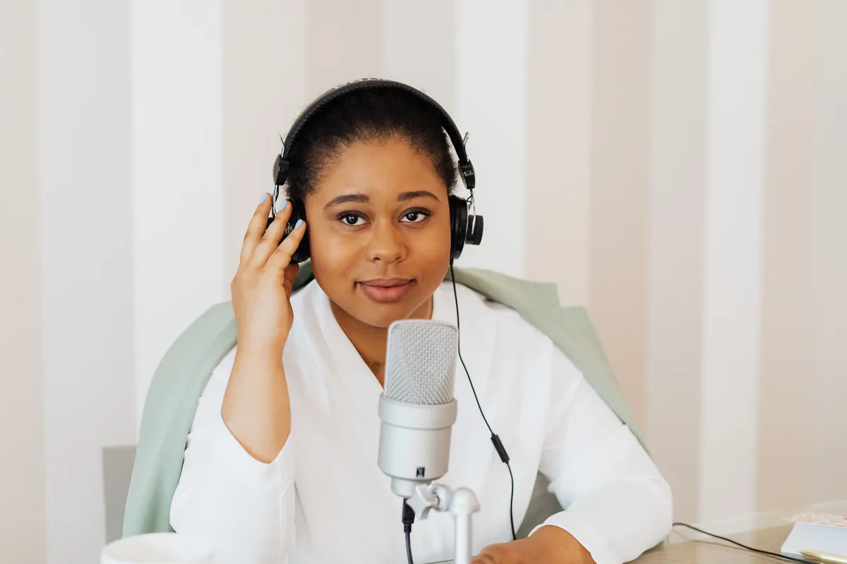 A podcaster in headphones speaking into a desk microphone in a bright room.